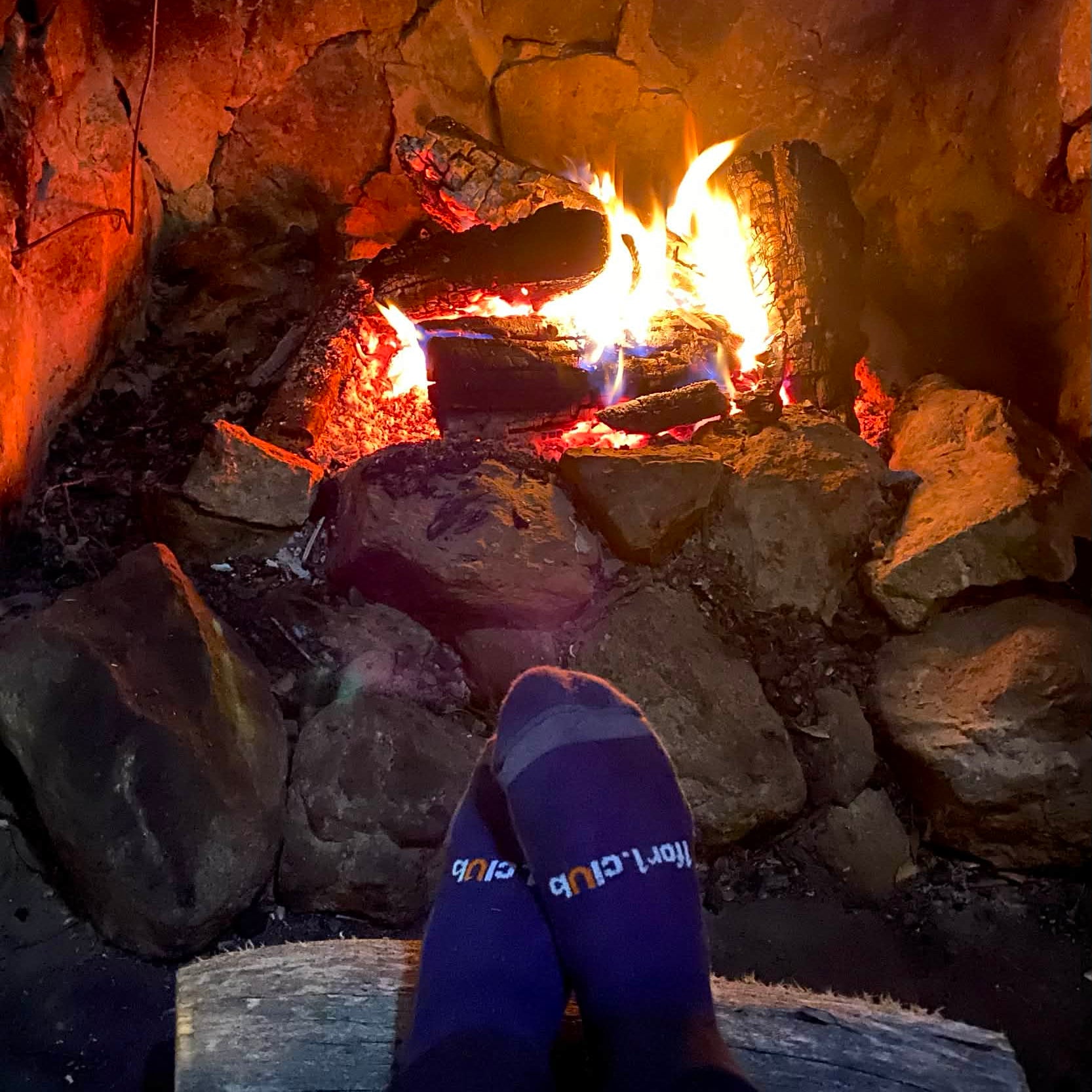 Person wearing blue socks with text sitting by a stone fire pit with flames.