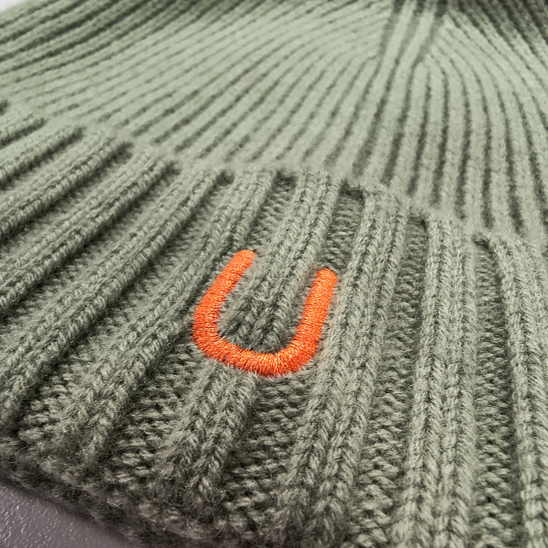 Close-up of front of 100% merino wool eucalyptus green ribbed knit beanie with bright orange embroidered U logo, showing fine texture and stitching detail